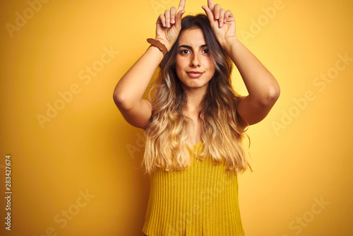 Young beautiful woman wearing t-shirt over yellow isolated background doing funny gesture with finger over head as bull horns