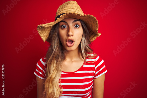 Young beautiful woman wearing summer hat over red isolated background scared in shock with a surprise face, afraid and excited with fear expression