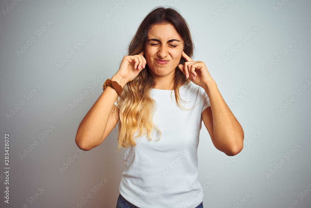 Fototapeta premium Young beautiful woman wearing casual white t-shirt over isolated background covering ears with fingers with annoyed expression for the noise of loud music. Deaf concept.