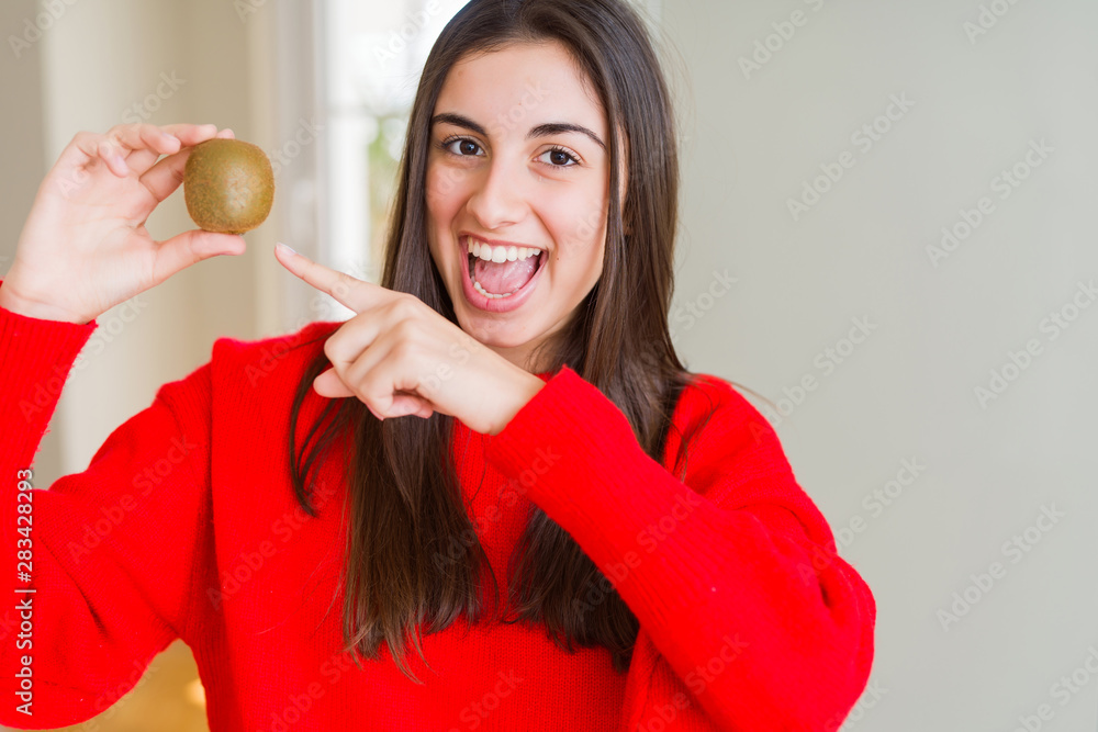 Beautiful young woman eating fresh green kiwi very happy pointing with hand and finger