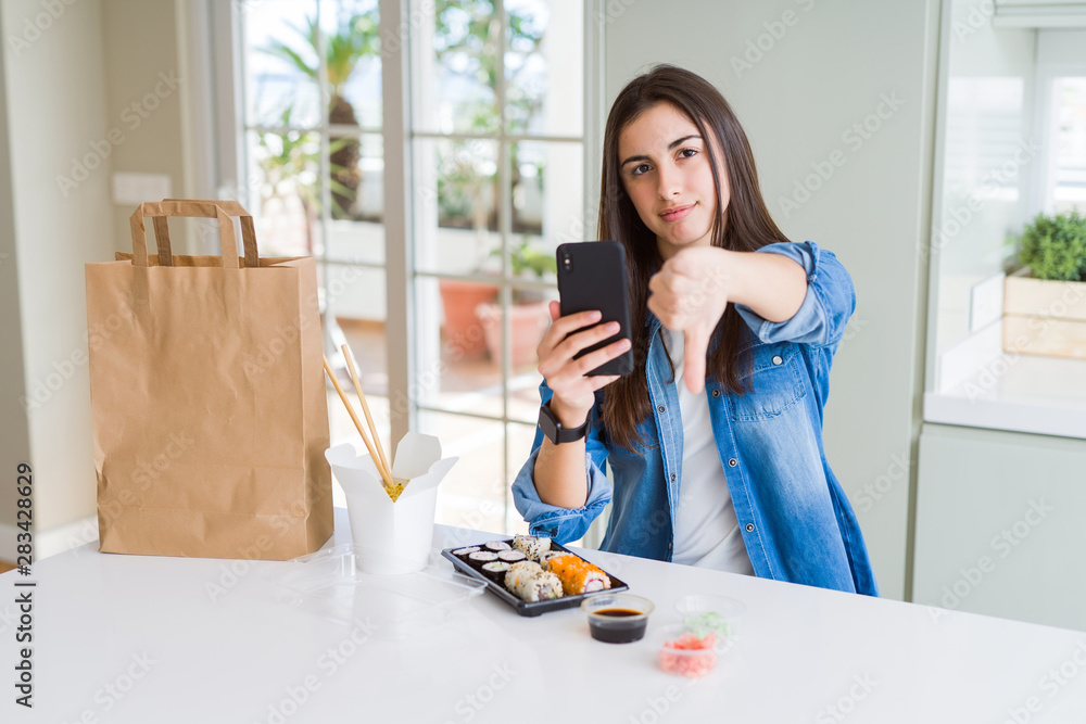 Beautiful young woman ordering food delivery from app using smartphone with angry face, negative sign showing dislike with thumbs down, rejection concept Beautiful young woman ordering food delivery from app using smartphone with angry face, negative sign showing dislike with thumbs down, rejection concept