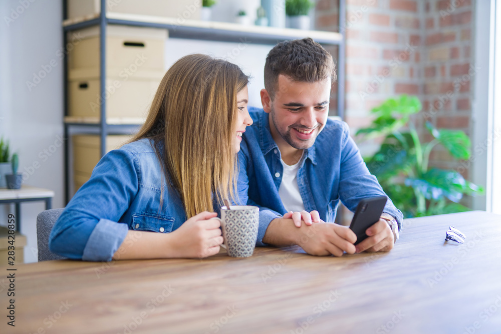 Young couple relaxing drinking a cup of coffee and using smartphone, sitting at new home with cardboard boxes behind them Young couple relaxing drinking a cup of coffee and using smartphone, sitting at new home with cardboard boxes behind them