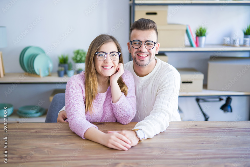 Young beautiful couple sitting on the table at home, hugging in love very happy for moving to new home with cardboard boxes behind them Young beautiful couple sitting on the table at home, hugging in love very happy for moving to new home with cardboard boxes behind them