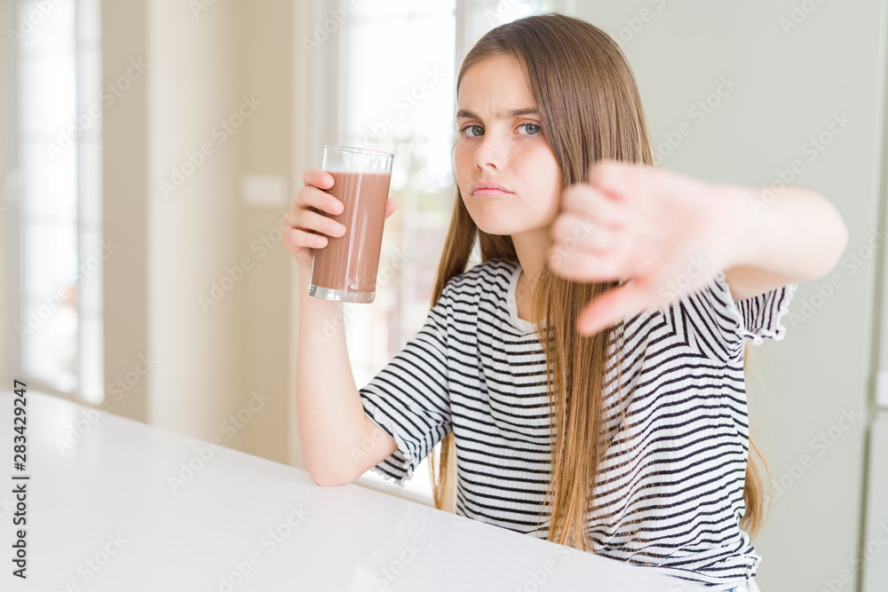 Beautiful young girl kid drinking fresh tasty chocolate milkshake as snack with angry face, negative sign showing dislike with thumbs down, rejection concept Beautiful young girl kid drinking fresh tasty chocolate milkshake as snack with angry face, negative sign showing dislike with thumbs down, rejection concept