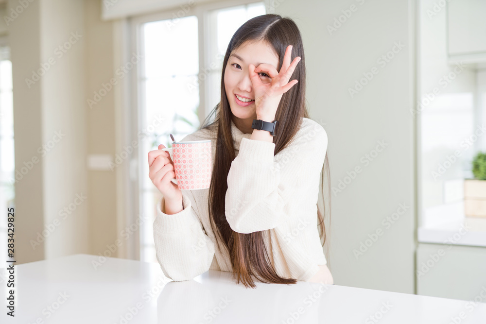 Beautiful Asian woman drinking a cup of coffee with happy face smiling doing ok sign with hand on eye looking through fingers Beautiful Asian woman drinking a cup of coffee with happy face smiling doing ok sign with hand on eye looking through fingers