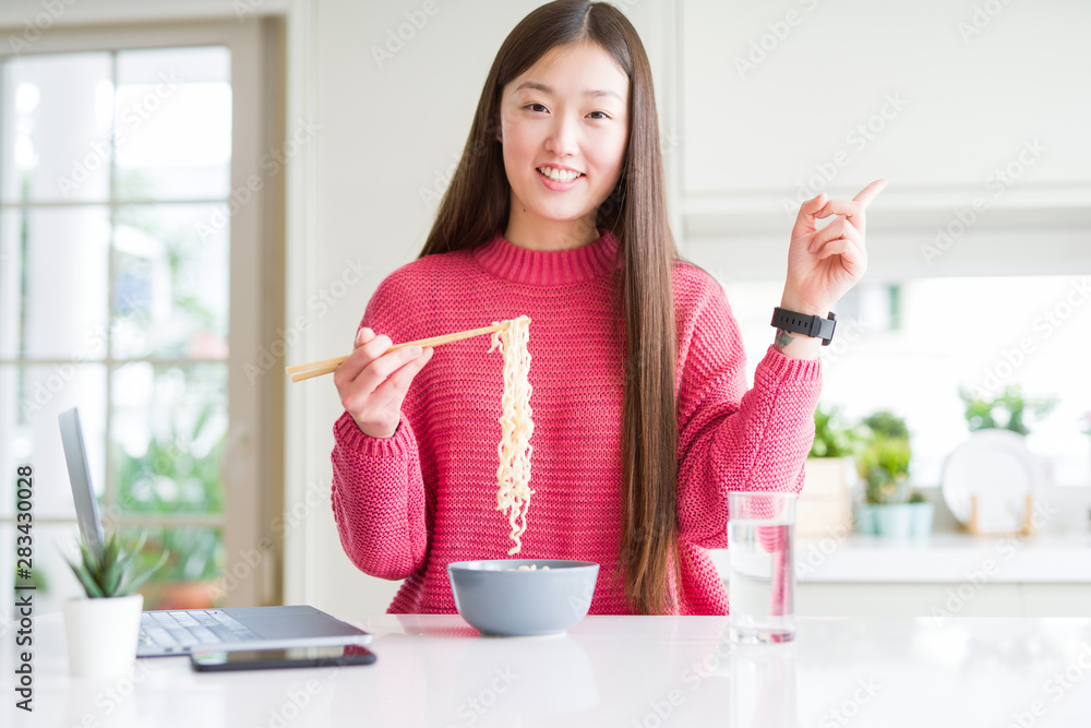 Beautiful Asian woman working using laptop and eating asian noodles very happy pointing with hand and finger to the side Beautiful Asian woman working using laptop and eating asian noodles very happy pointing with hand and finger to the side