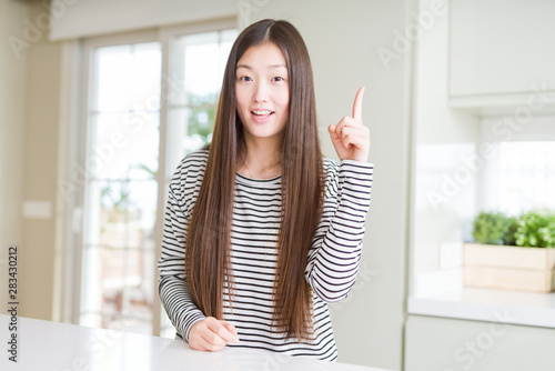 Beautiful Asian woman wearing stripes sweater pointing finger up with successful idea. Exited and happy. Number one.