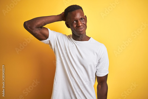 Young african american man wearing white t-shirt standing over isolated yellow background confuse and wonder about question. Uncertain with doubt, thinking with hand on head. Pensive concept.