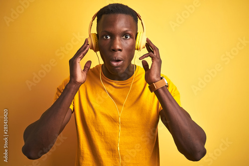 African american man listening to music using headphones over isolated yellow background scared in shock with a surprise face, afraid and excited with fear expression