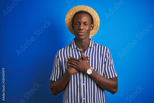 African american man wearing striped shirt and summer hat over isolated blue background smiling with hands on chest with closed eyes and grateful gesture on face. Health concept. © Krakenimages.com