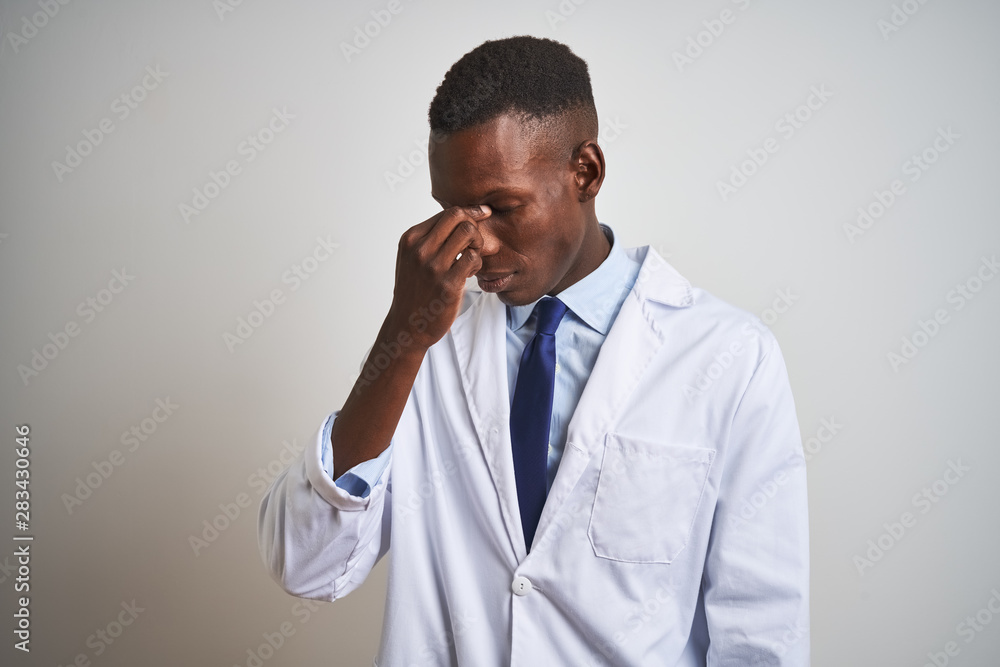 Young african american doctor man wearing coat standing over isolated white background tired rubbing nose and eyes feeling fatigue and headache. Stress and frustration concept. Young african american doctor man wearing coat standing over isolated white background tired rubbing nose and eyes feeling fatigue and headache. Stress and frustration concept.