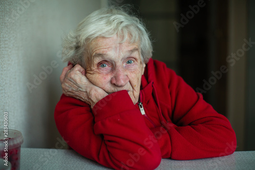 An old pensioner woman in her house, closeup portrait sitting at a table in a red jacket.