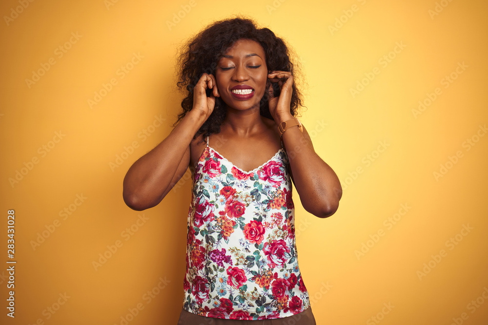 African american woman wearing floral summer t-shirt over isolated yellow background covering ears with fingers with annoyed expression for the noise of loud music. Deaf concept. African american woman wearing floral summer t-shirt over isolated yellow background covering ears with fingers with annoyed expression for the noise of loud music. Deaf concept.