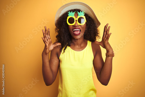 African american woman wearing funny pineapple sunglasses over isolated yellow background celebrating mad and crazy for success with arms raised and closed eyes screaming excited. Winner concept