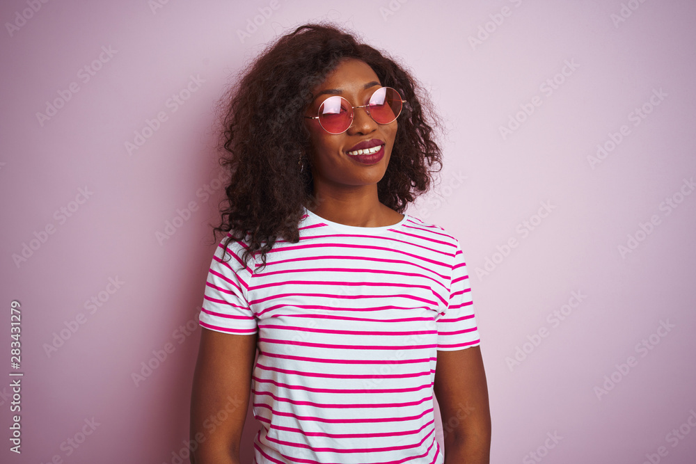 Young african american woman wearing t-shirt and sunglasses over isolated pink background looking away to side with smile on face, natural expression. Laughing confident.