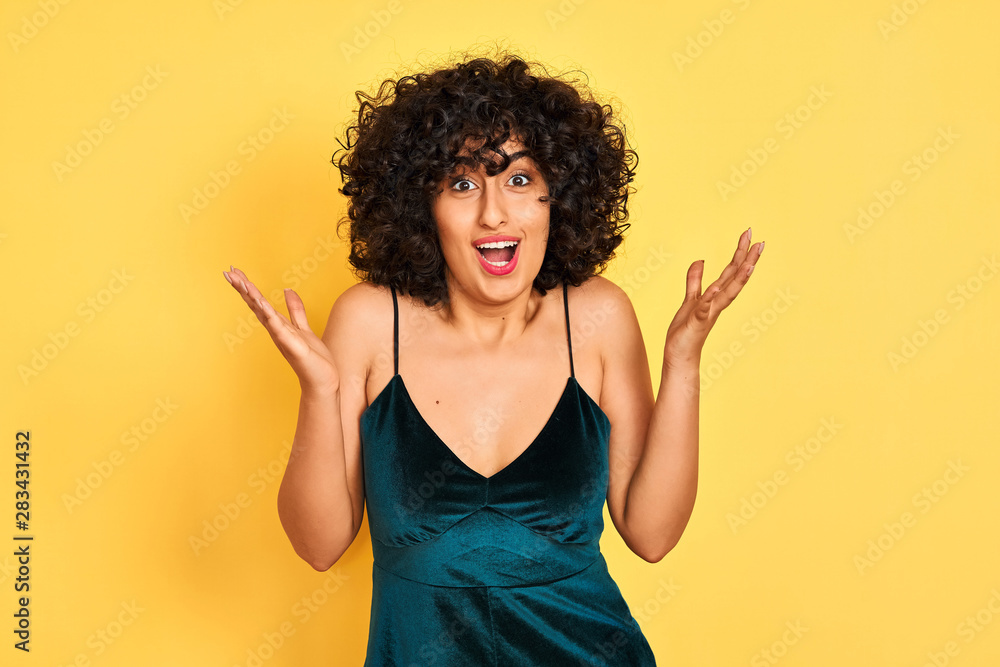 Young arab woman with curly hair wearing elegant dress over isolated yellow background celebrating crazy and amazed for success with arms raised and open eyes screaming excited. Winner concept Young arab woman with curly hair wearing elegant dress over isolated yellow background celebrating crazy and amazed for success with arms raised and open eyes screaming excited. Winner concept