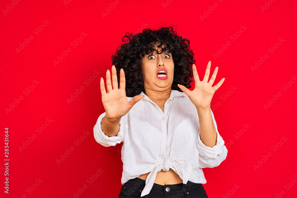 Young arab woman with curly hair wearing white casual shirt over isolated red background afraid and terrified with fear expression stop gesture with hands, shouting in shock. Panic concept. Young arab woman with curly hair wearing white casual shirt over isolated red background afraid and terrified with fear expression stop gesture with hands, shouting in shock. Panic concept.