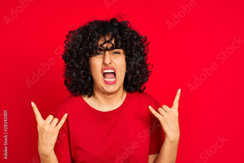 Young arab woman with curly hair wearing casual t-shirt over isolated red background shouting with crazy expression doing rock symbol with hands up. Music star. Heavy concept.