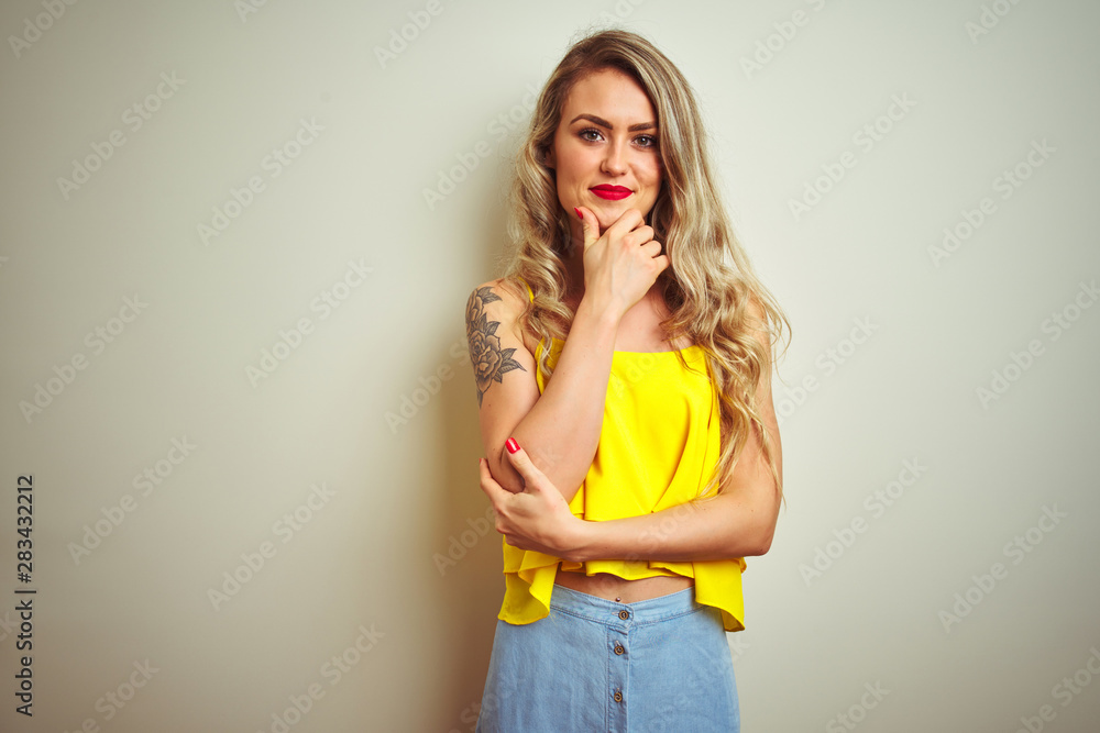 Young beautiful woman wearing yellow t-shirt standing over white isolated background looking confident at the camera with smile with crossed arms and hand raised on chin. Thinking positive. Young beautiful woman wearing yellow t-shirt standing over white isolated background looking confident at the camera with smile with crossed arms and hand raised on chin. Thinking positive.