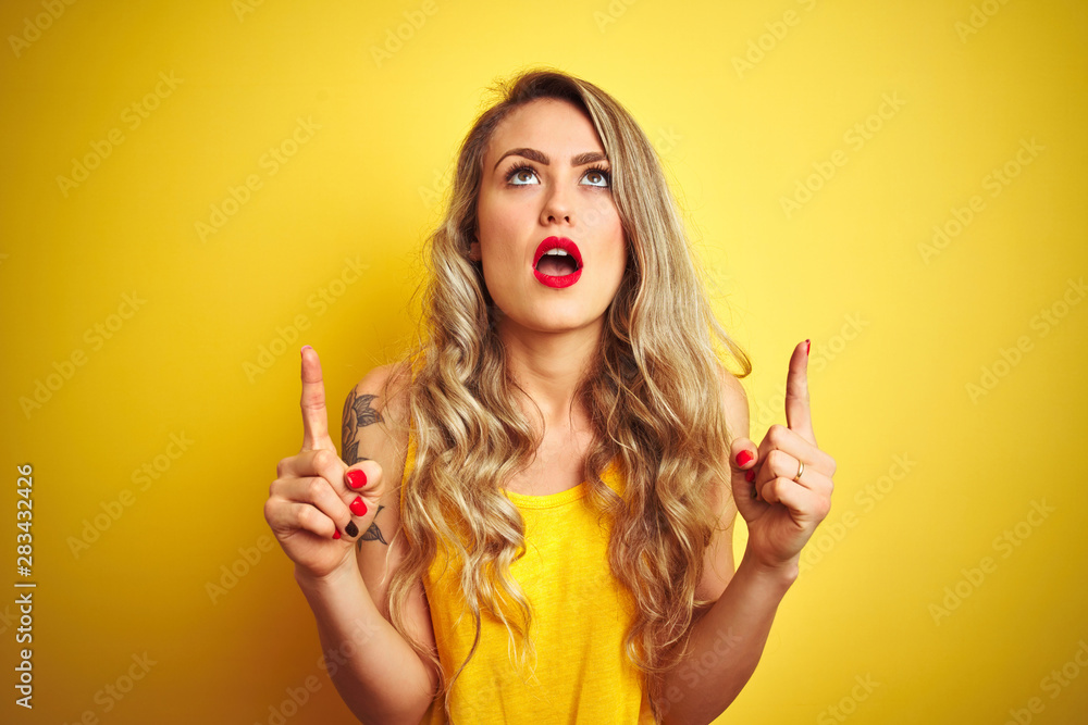 Obraz premium Young beautiful woman wearing t-shirt standing over yellow isolated background amazed and surprised looking up and pointing with fingers and raised arms.