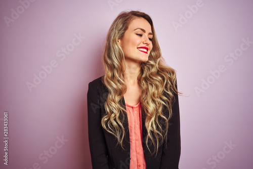 Young beautiful business woman wearing elegant jacket standing over pink isolated background looking away to side with smile on face, natural expression. Laughing confident.