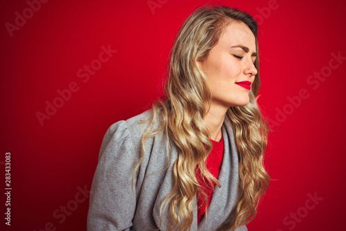 Young beautiful business woman wearing elegant jacket standing over red isolated background with hand on stomach because nausea, painful disease feeling unwell. Ache concept.