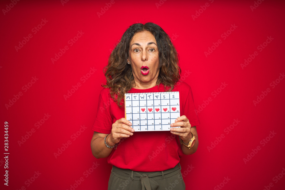 Middle age senior woman holding menstruation calendar over red isolated background scared in shock with a surprise face, afraid and excited with fear expression Middle age senior woman holding menstruation calendar over red isolated background scared in shock with a surprise face, afraid and excited with fear expression