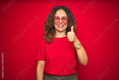 Middle age senior woman wearing cute heart shaped glasses over red isolated background doing happy thumbs up gesture with hand. Approving expression looking at the camera with showing success.
