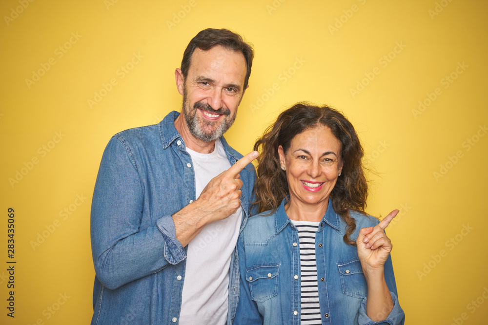 Beautiful middle age couple together wearing denim shirt over isolated yellow background cheerful with a smile of face pointing with hand and finger up to the side with happy and natural expression