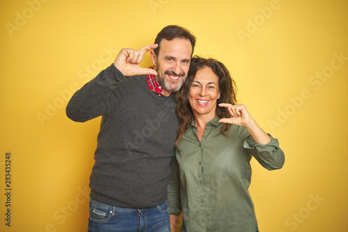 Beautiful middle age couple over isolated yellow background smiling and confident gesturing with hand doing small size sign with fingers looking and the camera. Measure concept.