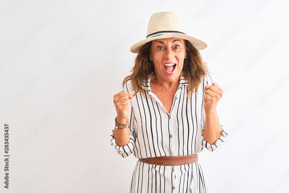 Middle age businesswoman wearing striped dress and hat over isolated white background celebrating surprised and amazed for success with arms raised and open eyes. Winner concept. Middle age businesswoman wearing striped dress and hat over isolated white background celebrating surprised and amazed for success with arms raised and open eyes. Winner concept.