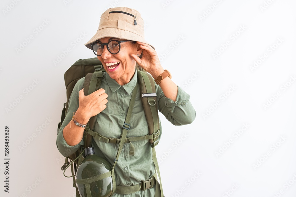 Middle age hiker woman wearing backpack canteen hat glasses over isolated white background smiling with hand over ear listening an hearing to rumor or gossip. Deafness concept. Middle age hiker woman wearing backpack canteen hat glasses over isolated white background smiling with hand over ear listening an hearing to rumor or gossip. Deafness concept.