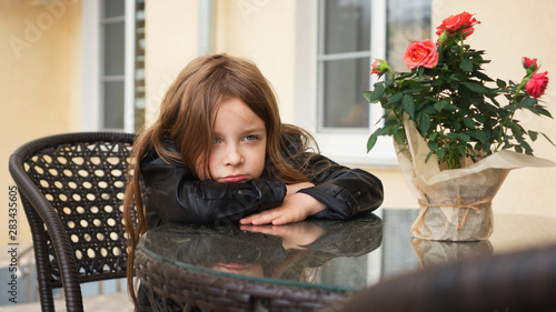 Fashionable little schoolgirl sitting at table of summer terrace of restaurant. She's upset about waiting for order. photo