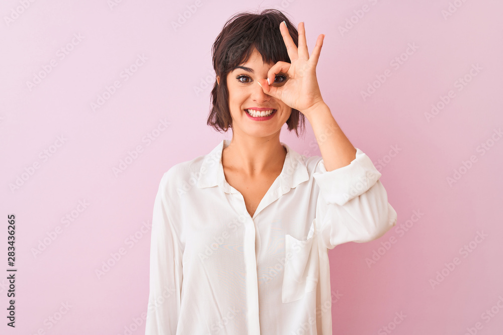 Young beautiful woman wearing white shirt standing over isolated pink background doing ok gesture with hand smiling, eye looking through fingers with happy face. Young beautiful woman wearing white shirt standing over isolated pink background doing ok gesture with hand smiling, eye looking through fingers with happy face.