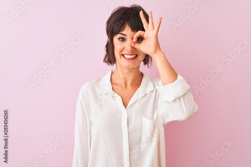 Young beautiful woman wearing white shirt standing over isolated pink background doing ok gesture with hand smiling, eye looking through fingers with happy face. © Krakenimages.com