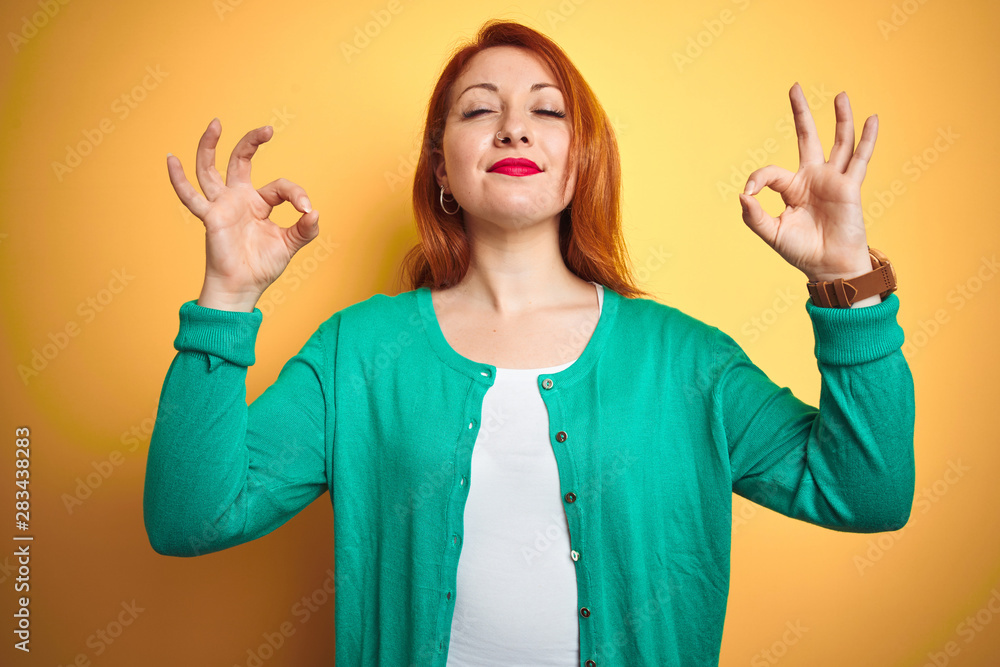 Youg beautiful redhead woman wearing winter green sweater over isolated yellow background relax and smiling with eyes closed doing meditation gesture with fingers. Yoga concept.