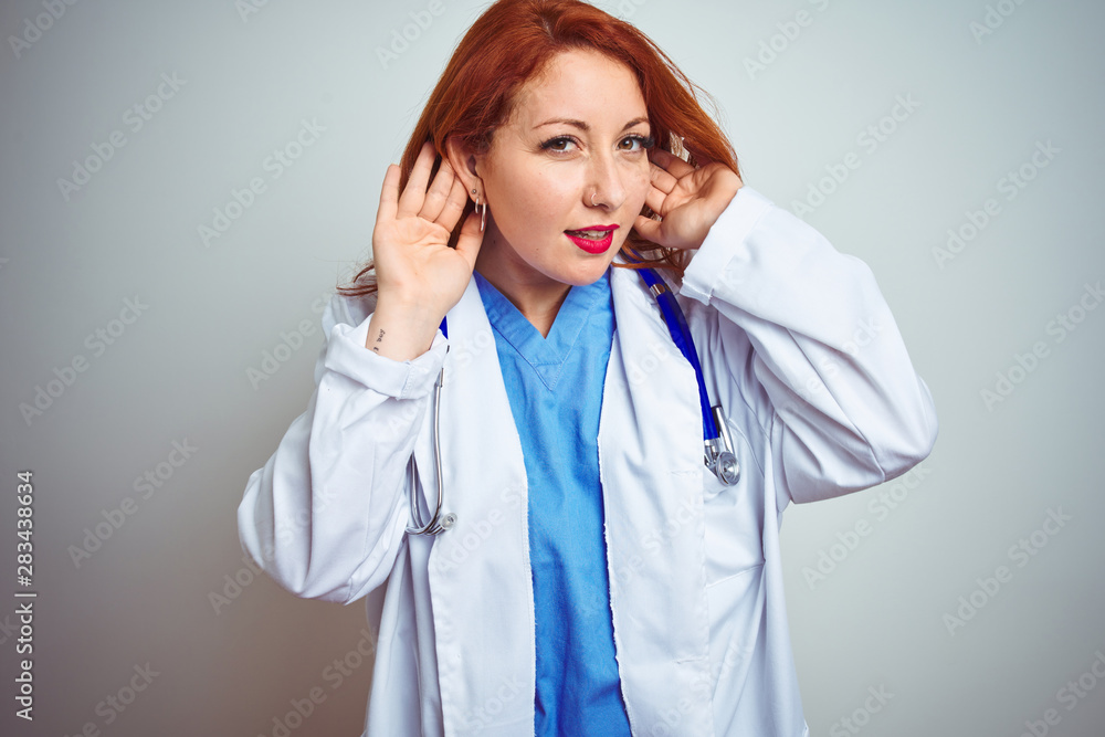 Young redhead doctor woman using stethoscope over white isolated background Trying to hear both hands on ear gesture, curious for gossip. Hearing problem, deaf Young redhead doctor woman using stethoscope over white isolated background Trying to hear both hands on ear gesture, curious for gossip. Hearing problem, deaf