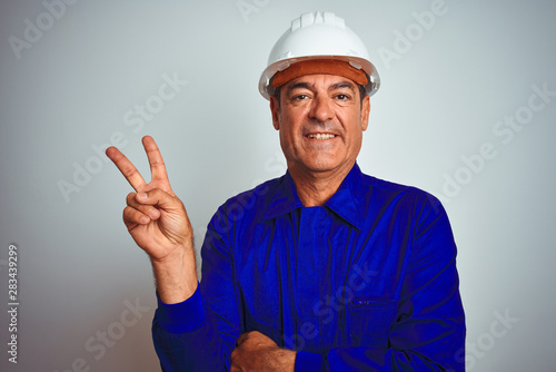 Handsome middle age worker man wearing uniform and helmet over isolated white background smiling with happy face winking at the camera doing victory sign. Number two.