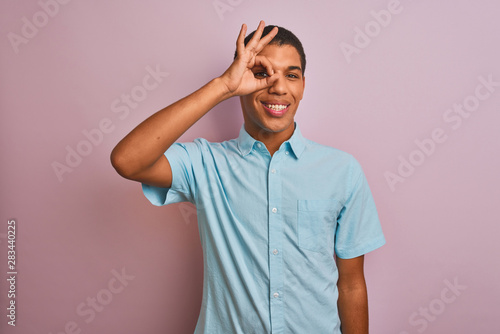 Young handsome arab man wearing blue shirt standing over isolated pink background doing ok gesture with hand smiling, eye looking through fingers with happy face.