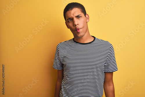 Young handsome arab man wearing navy striped t-shirt over isolated yellow background making fish face with lips, crazy and comical gesture. Funny expression.