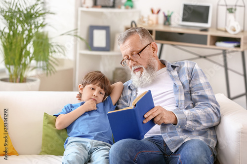 Cute little boy reading book with grandfather at home