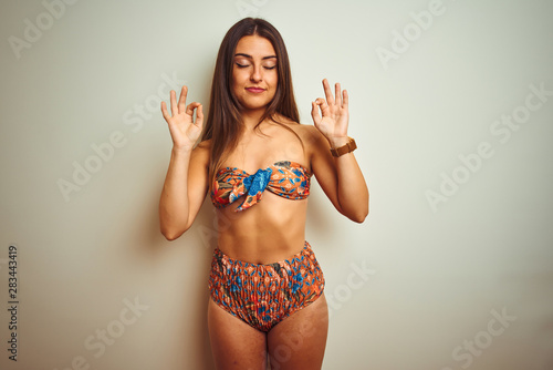 Young beautiful woman on vacation wearing bikini standing over isolated white background relax and smiling with eyes closed doing meditation gesture with fingers. Yoga concept.
