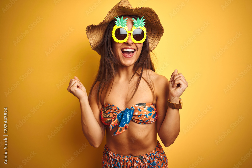 Fototapeta premium Woman on vacation wearing bikini and pineapple sunglasses over isolated yellow background celebrating surprised and amazed for success with arms raised and open eyes. Winner concept.