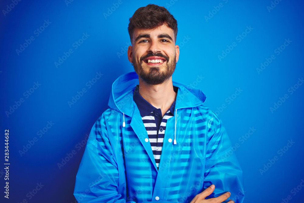 Young handsome man wearing rain coat standing over isolated blue background happy face smiling with crossed arms looking at the camera. Positive person.