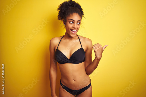 African american woman on vacation wearing bikini standing over isolated yellow background smiling with happy face looking and pointing to the side with thumb up. © Krakenimages.com