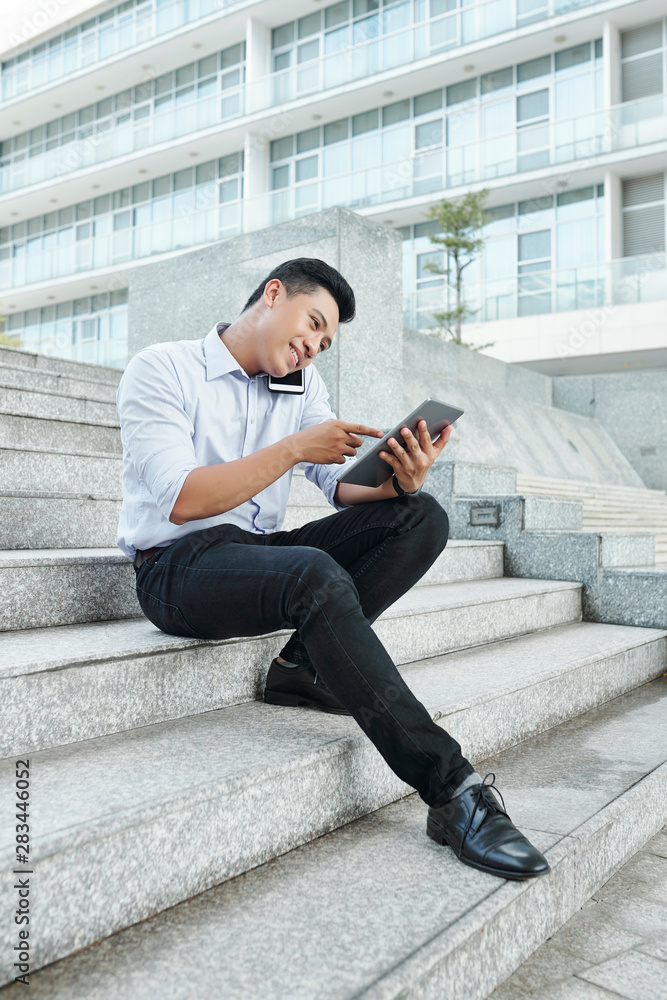 Smiling young Asian businessman sitting on steps, calling on phone and reading information on digital tablet