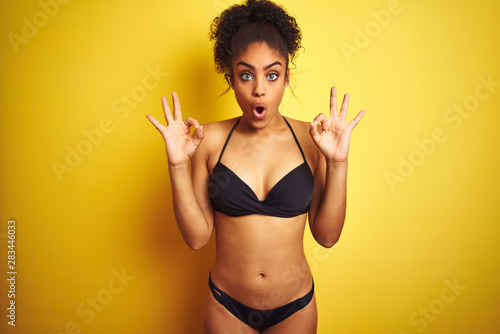 African american woman on vacation wearing bikini standing over isolated yellow background looking surprised and shocked doing ok approval symbol with fingers. Crazy expression