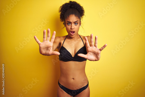 African american woman on vacation wearing bikini standing over isolated yellow background doing stop gesture with hands palms, angry and frustration expression
