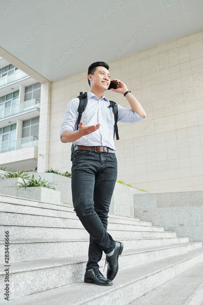 Handsome young Asian university student with backpack standing on campus and calling on phone Handsome young Asian university student with backpack standing on campus and calling on phone