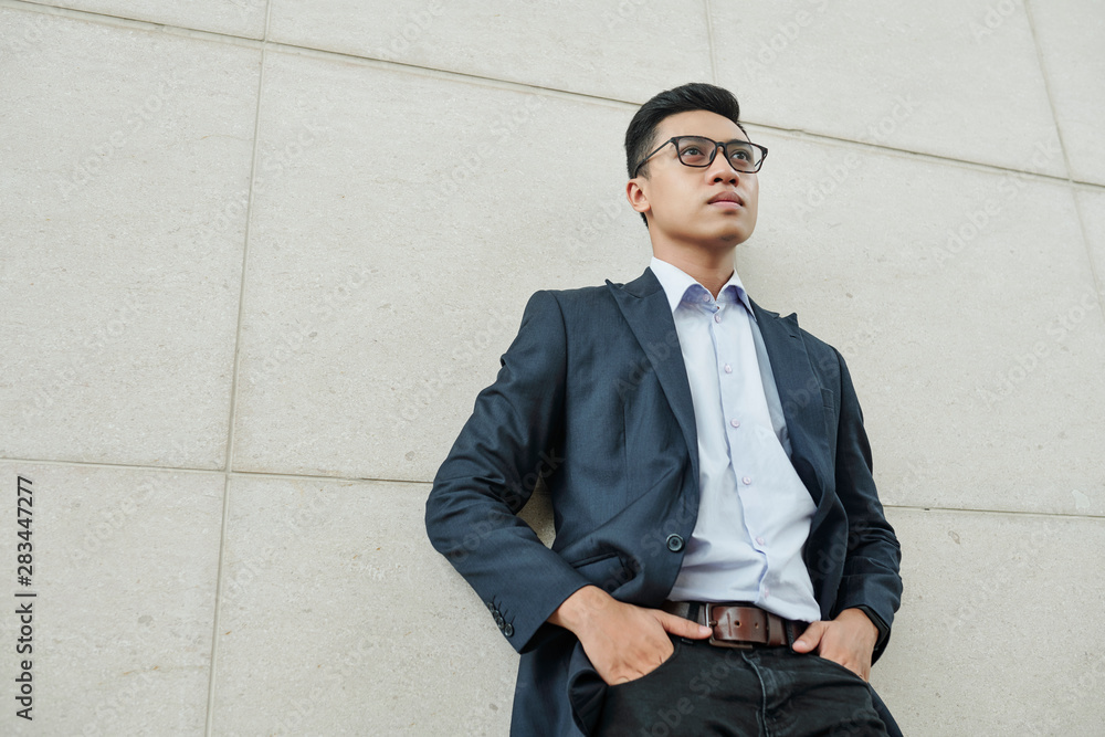Confident young Vietnamese businessman in glasses putting hands in pockets when standing at building wall and looking in front of him Confident young Vietnamese businessman in glasses putting hands in pockets when standing at building wall and looking in front of him